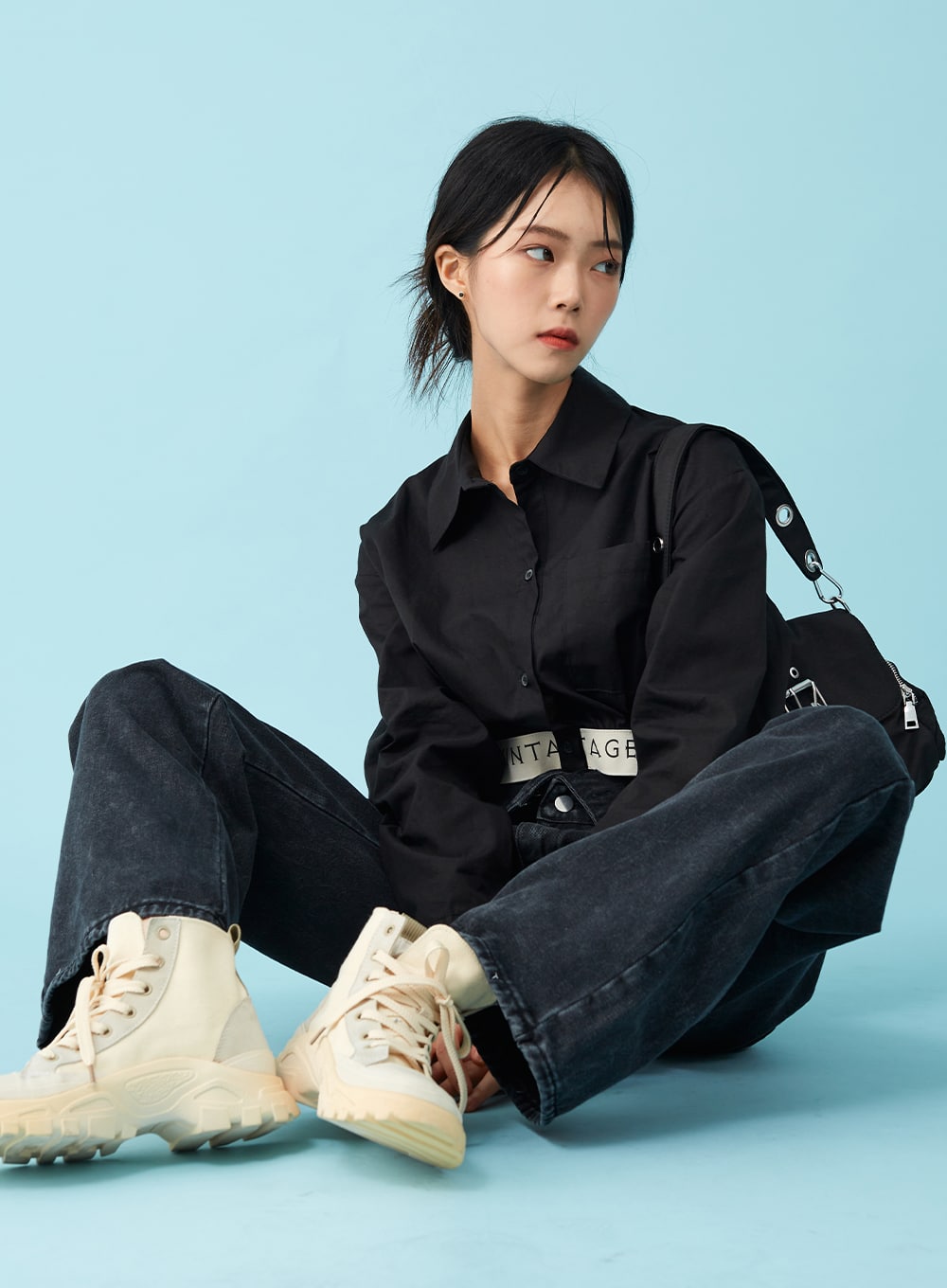 Four Buttoned Open Black Jeans CU8 in washed black, seated three-quarter view on model looking aside, wide washed-denim legs splayed, logo belt at open waistband, black crop shirt and crossbody bag.