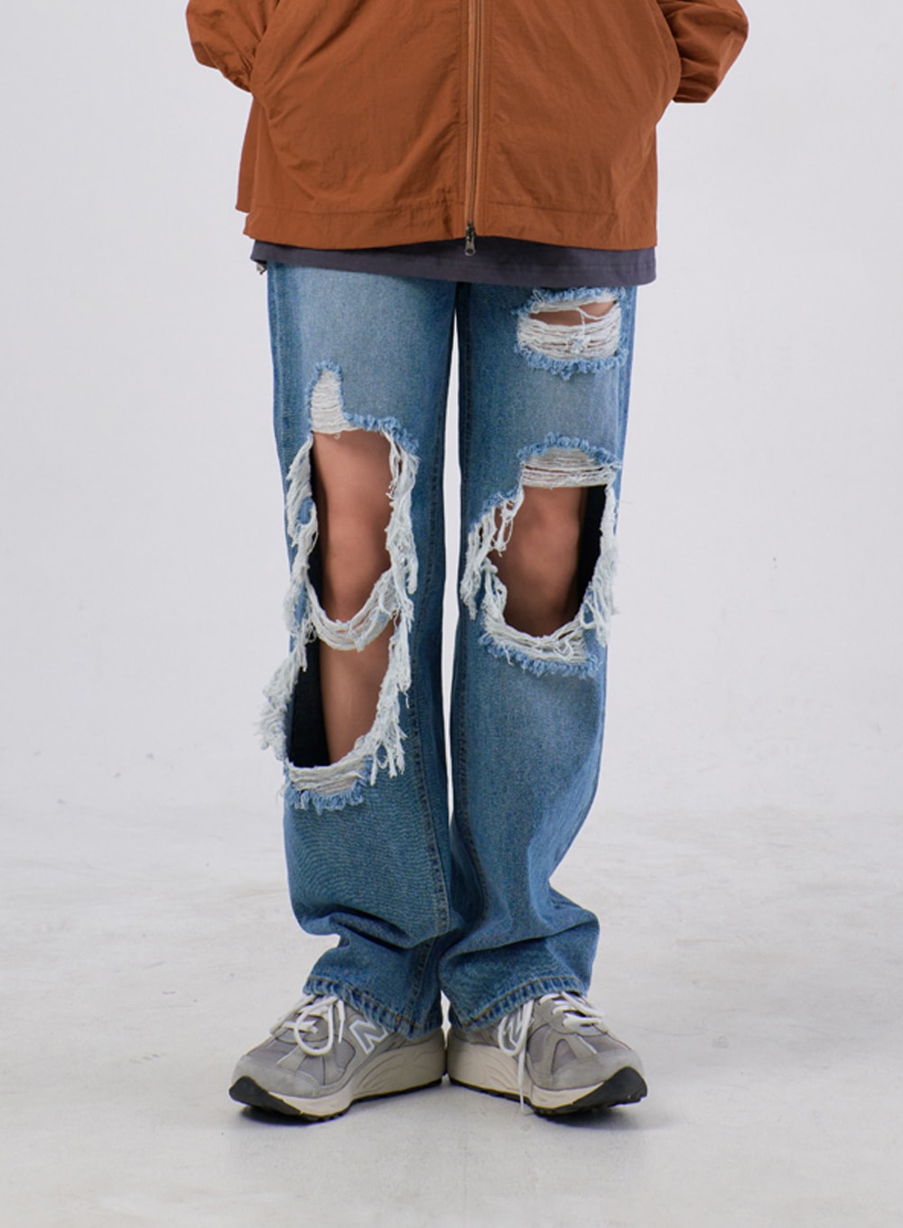 Ripped Knee Jeans IA305 in medium blue, front lower-body close-up on model, showcasing oversized destroyed knee openings with long frayed threads, worn under brown windbreaker hem.