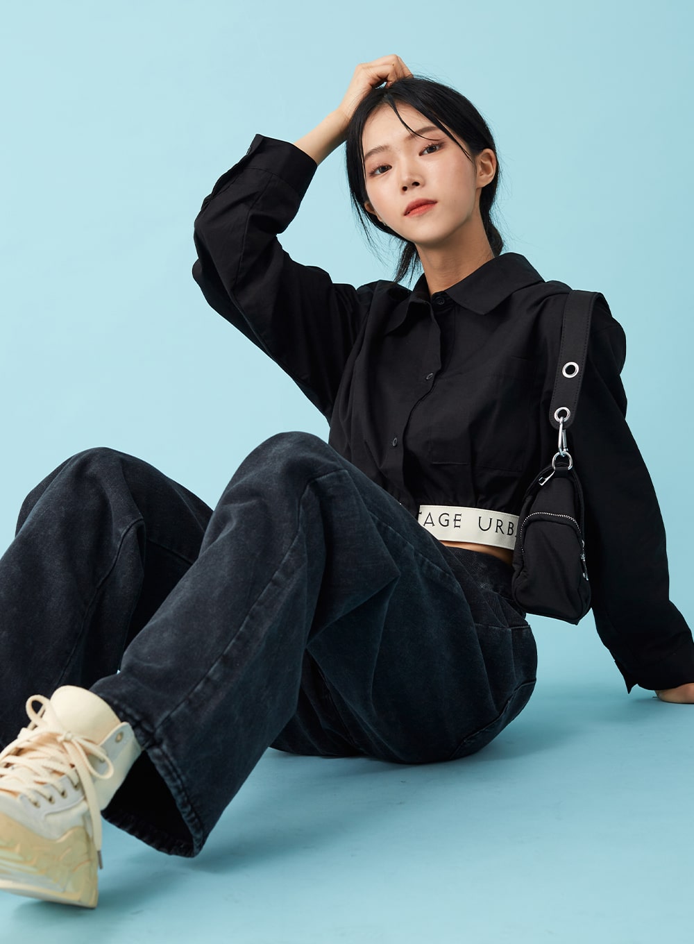 Four Buttoned Open Black Jeans CU8 in washed black, seated close editorial pose on model, wide-leg denim with open four-button waistband and logo belt, black crop shirt, crossbody bag, chunky sneakers.