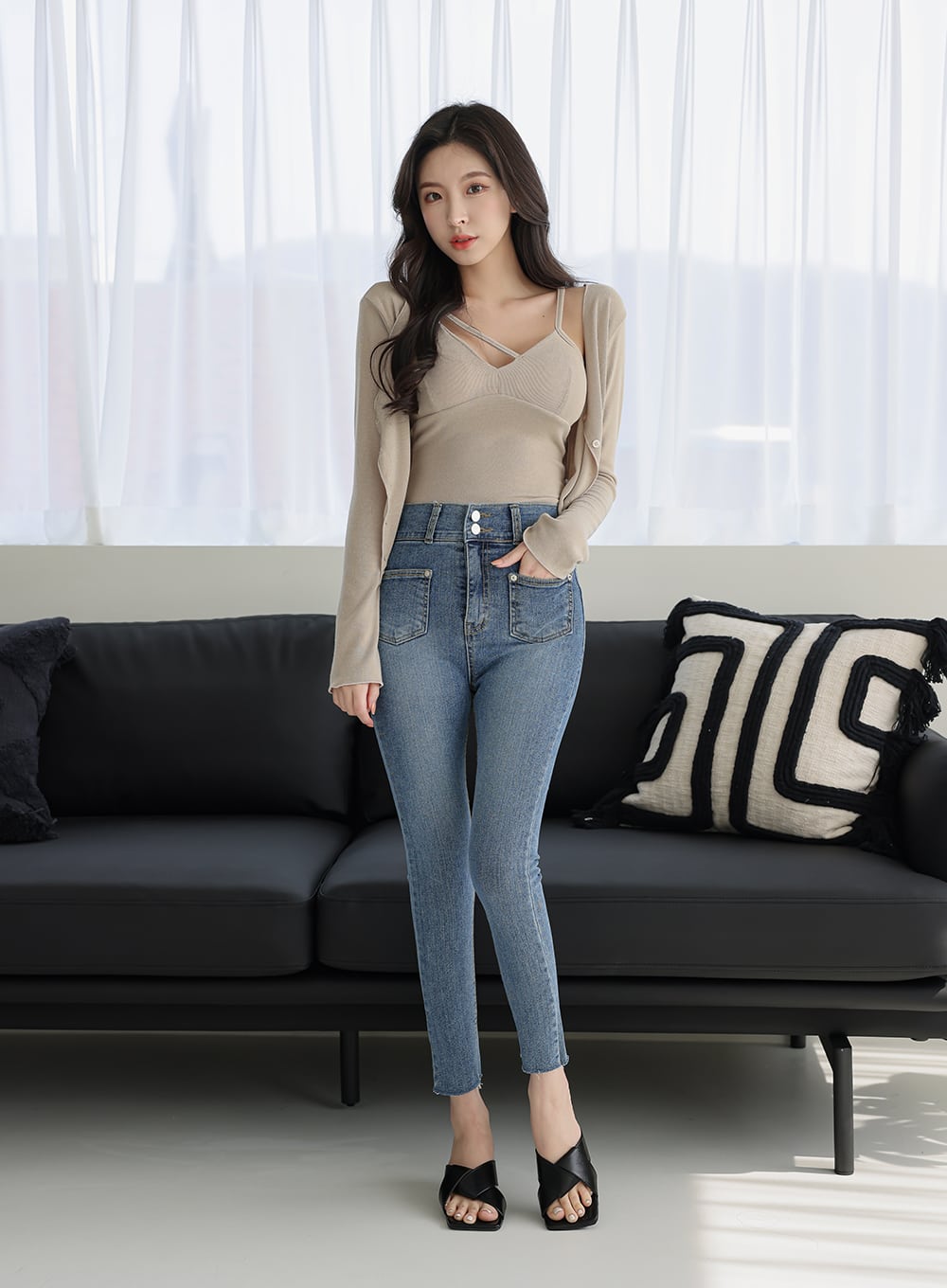 Double Button Raw Hem Skinny Jeans IA11 in mid blue denim, front full-body shot on model standing by dark sofa, high-rise waist with double buttons, skinny fit, paired with beige knit top and black mules.
