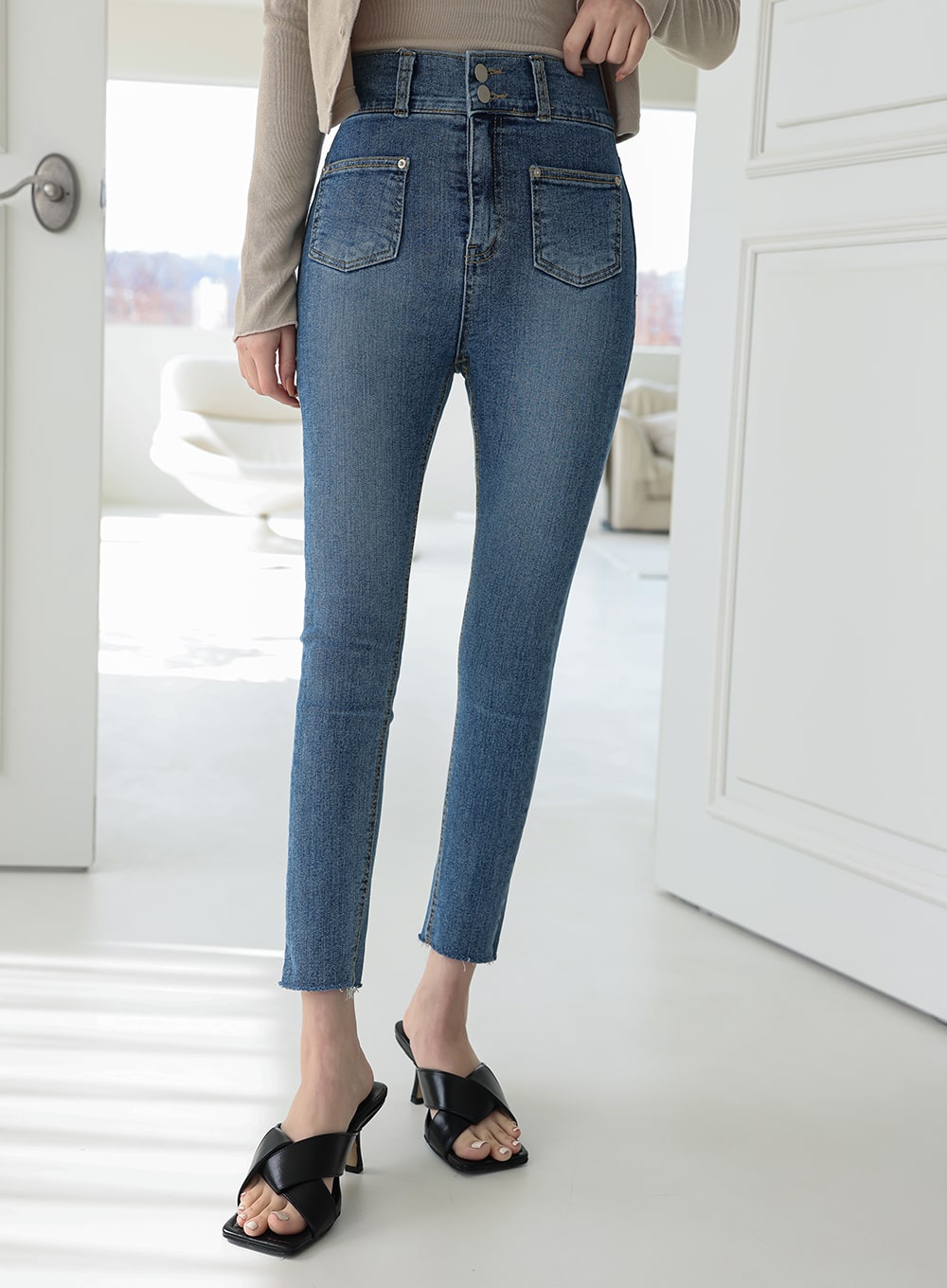 Double Button Raw Hem Skinny Jeans IA11 in mid blue denim, front lower-body shot on model, displaying slim leg silhouette, front patch pockets, and raw frayed hem at ankle with black mule sandals.