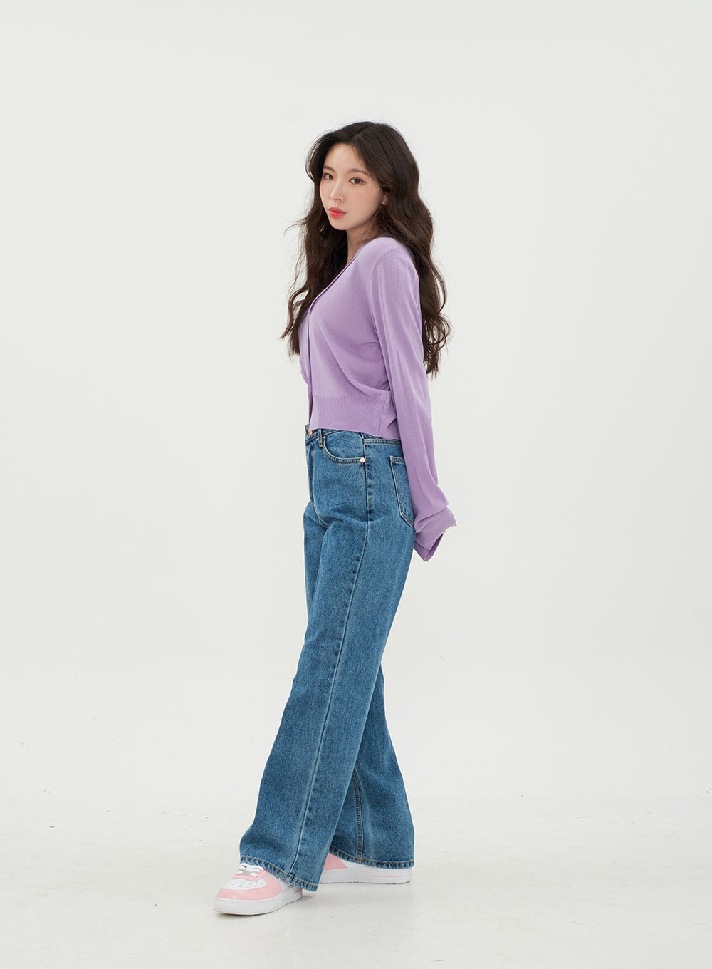 Semi Wide Jeans BM21 in mid-blue denim, three-quarter side view on model glancing back, wide-fit long denim with side pocket detail visible, styled with lavender cardigan and pink sneakers.