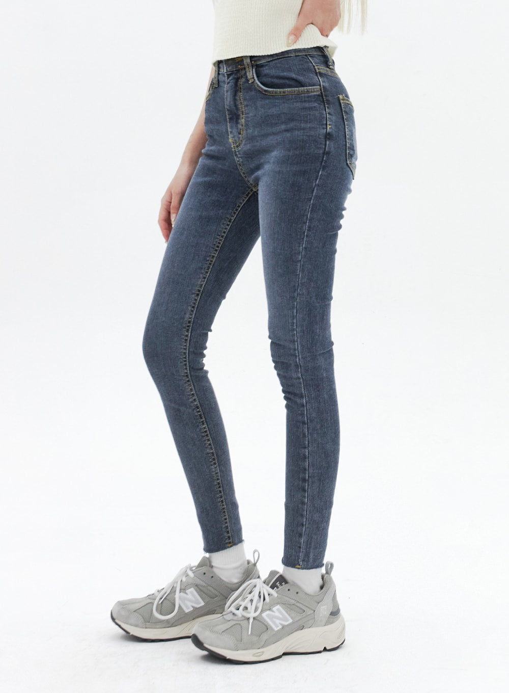 Skinny Ankle Jeans BA321 in dark blue denim, side view on model, high-rise fit hugging hips and thighs, tapered ankle hem, paired with cream ribbed top and grey New Balance sneakers.