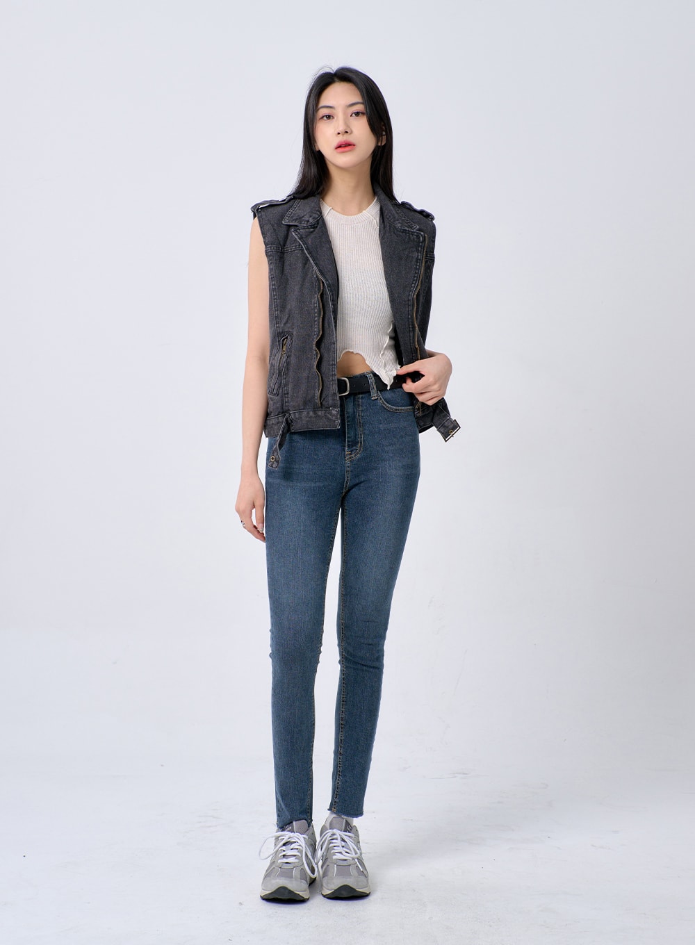 Skinny Ankle Jeans BA321 in dark blue denim, full-body front shot on model, high-rise skinny silhouette with black belt, styled with grey denim vest and cream ribbed crop top, grey sneakers.
