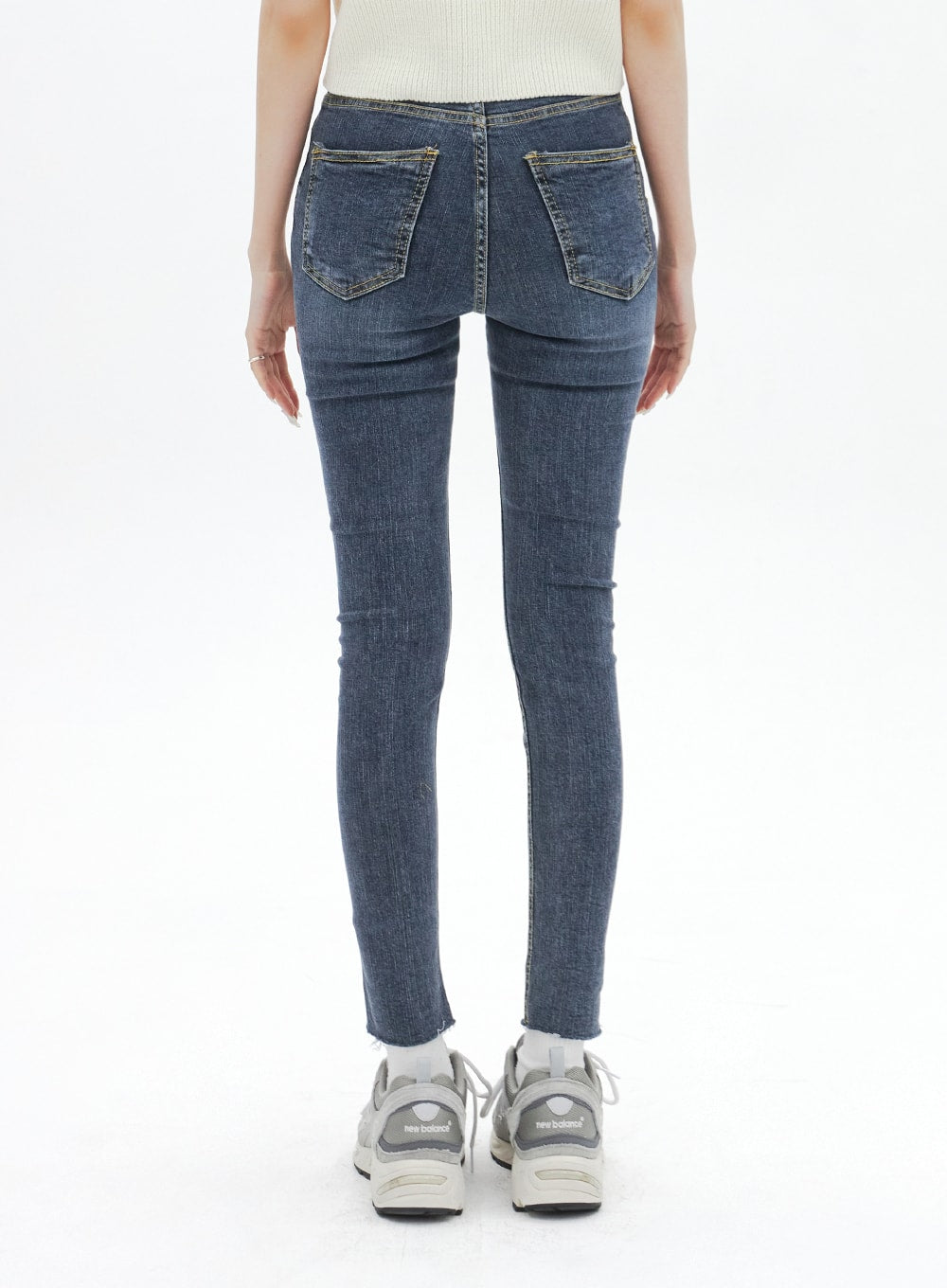Skinny Ankle Jeans BA321 in dark blue denim, back view on model, dual back patch pockets with gold stitching, slim leg tapering to raw ankle hem, worn with grey sneakers.