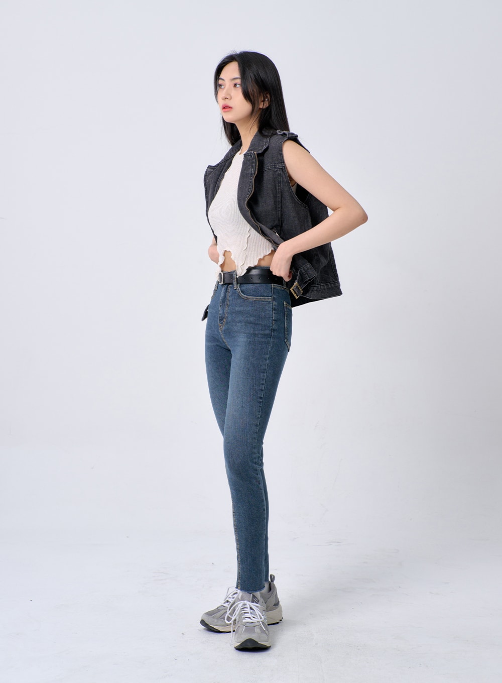 Skinny Ankle Jeans BA321 in dark blue denim, full-body side shot on model, high-rise slim leg tapering to ankle, black belt at waist, layered with charcoal denim vest and white ribbed crop top.